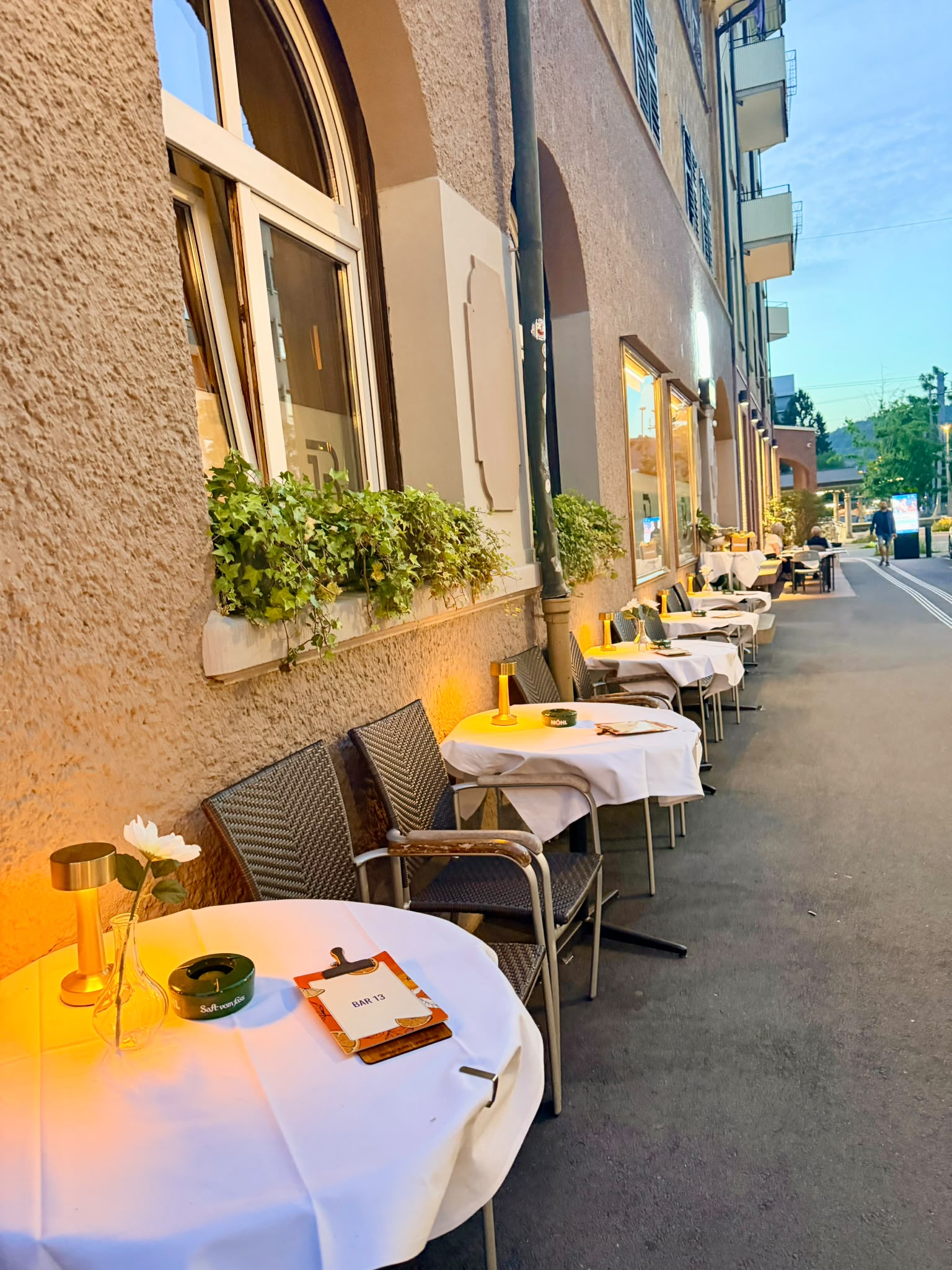 Terrasse der B13 Bar an der Bahnhofstrasse bei Dämmerung mit gedeckten Tischen und warmen Tischlampen
