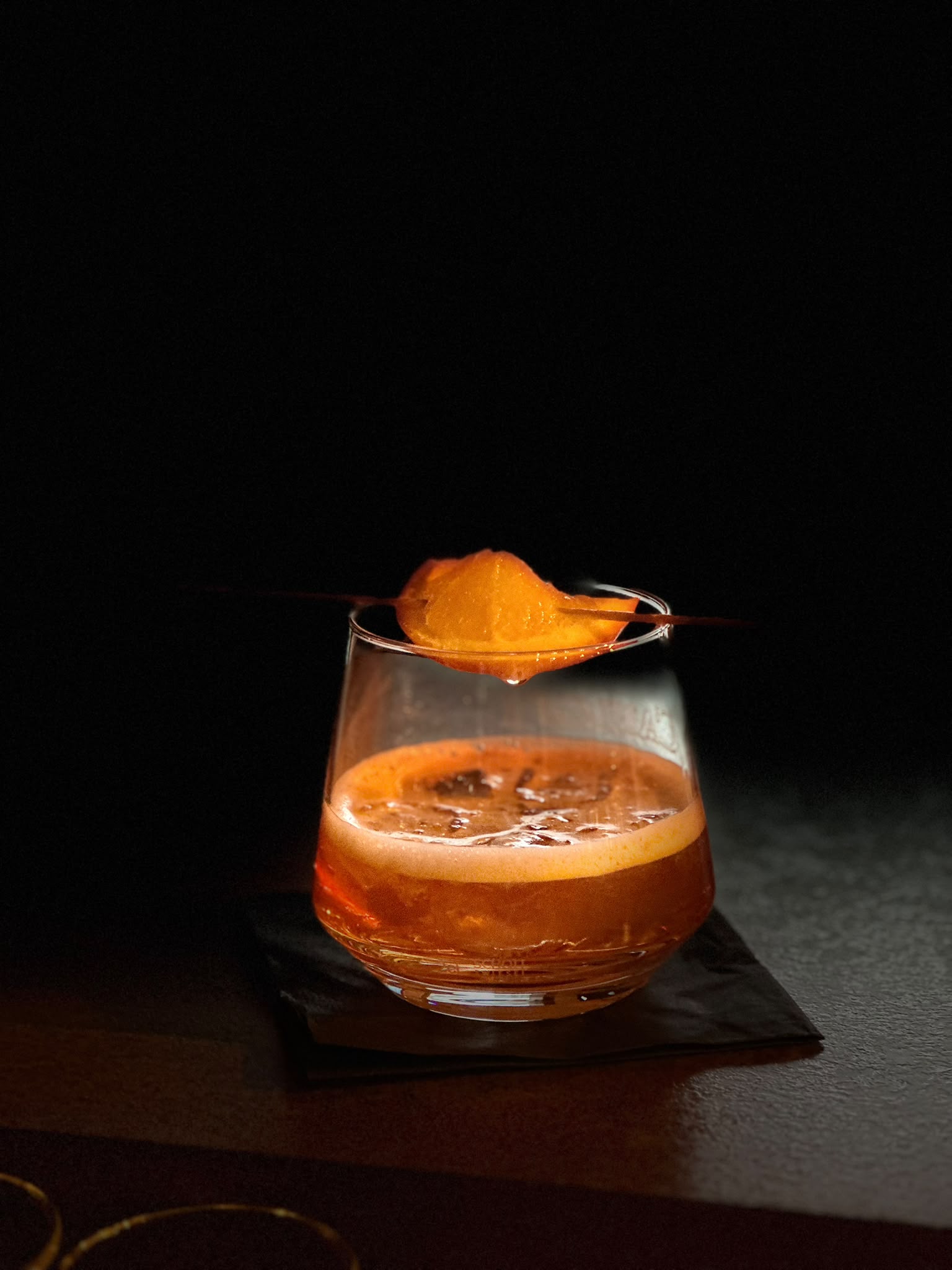Dunkler Signature-Cocktail mit Orangenzeste im warmen Spotlight